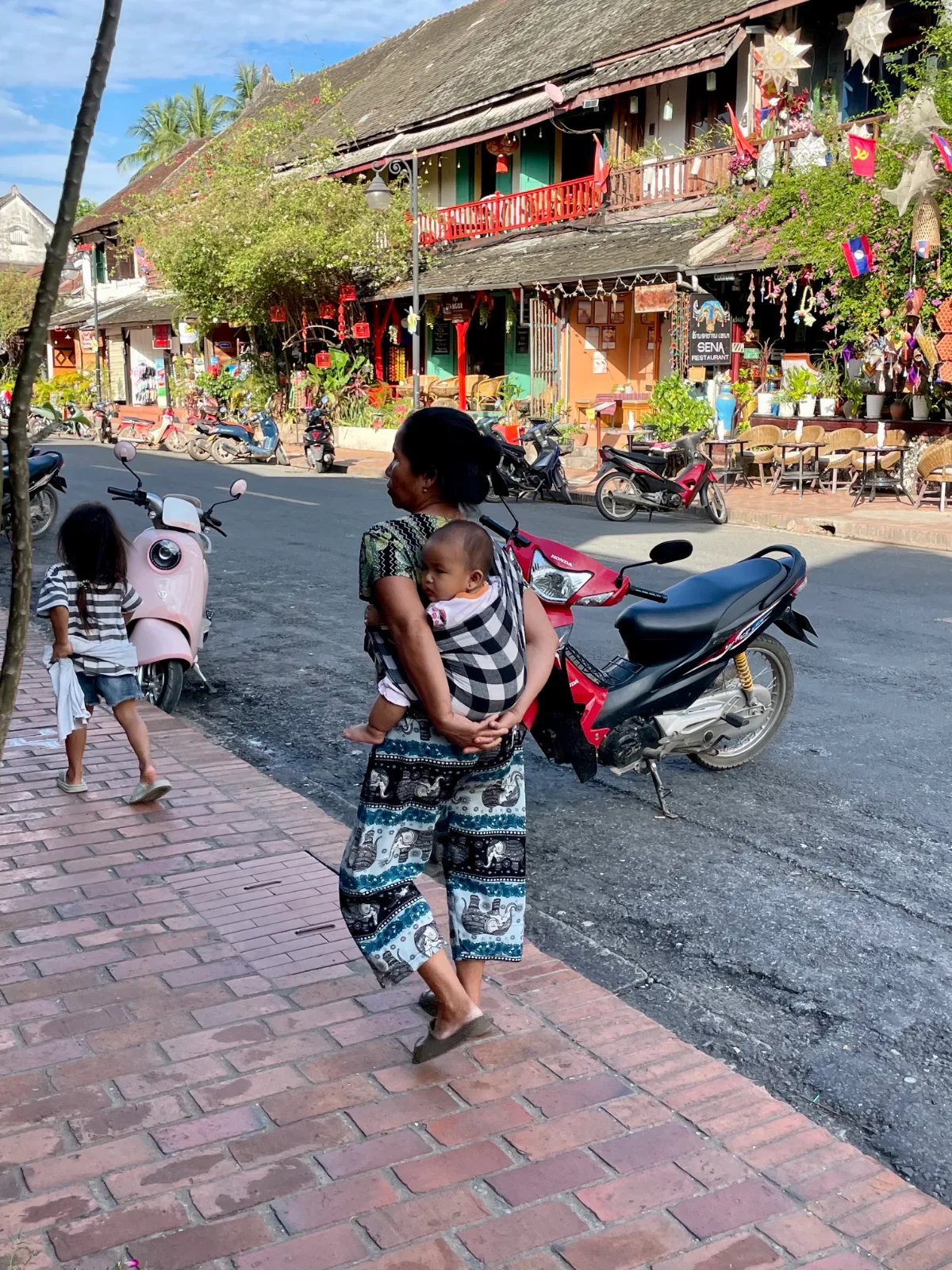 A woman with a baby on her back and a young girl walking down the street in Laos